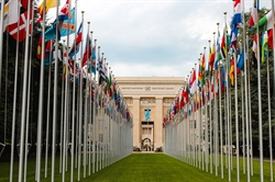 [ai] A row of flags from various countries displayed in front of the United Nations building in Geneva, framed by green grass and trees under a cloudy sky.